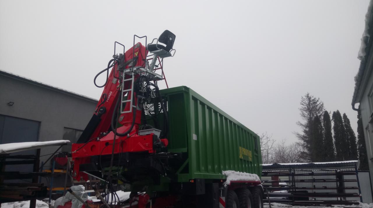 Czerwony żuraw leśny z siedziskiem na platformie, zamontowany na zielonej przyczepie Pronar, widoczny na tle szarego nieba i budynków z elementami śniegu.