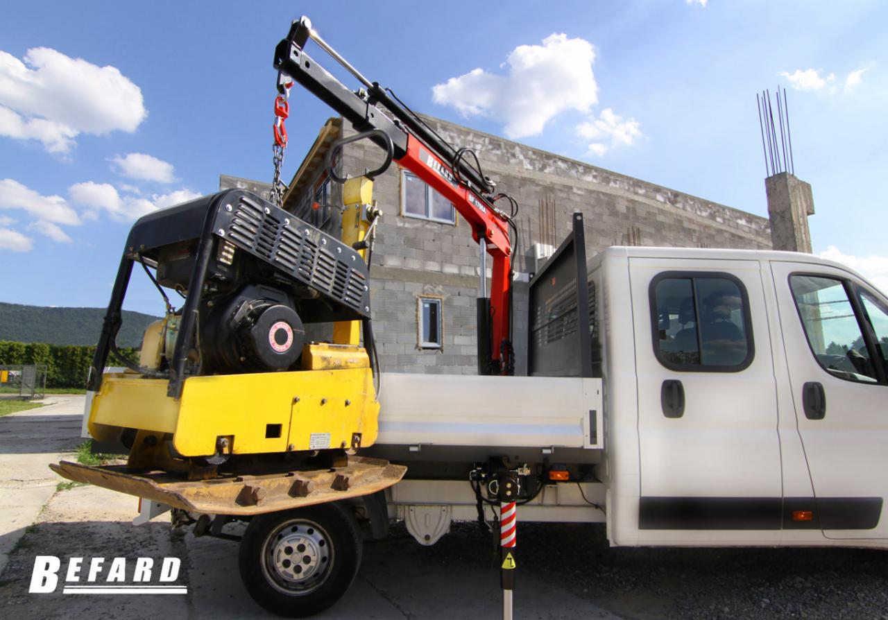 Żółta zagęszczarka gruntu podnoszona czerwoną mini żurawiem zamontowanym na białej platformie samochodu dostawczego, na tle budowanego budynku z szarych pustaków.