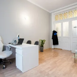 Jasne biuro: białe biurko z laptopem, krzesło obrotowe z białej skóry, toga prawnicza wisząca na drzwiach, podłoga z paneli.