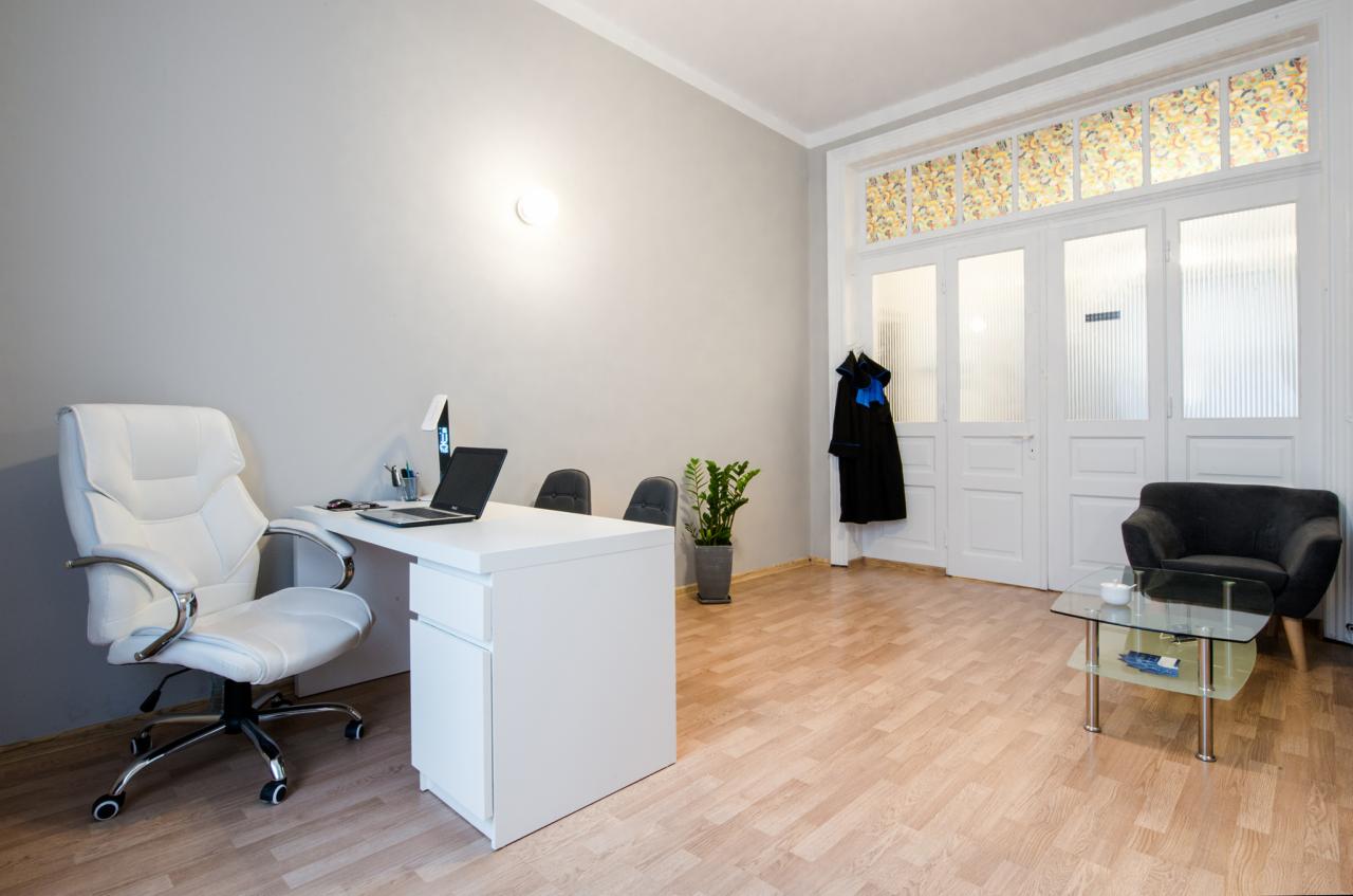 Jasne biuro: białe biurko z laptopem, krzesło obrotowe z białej skóry, toga prawnicza wisząca na drzwiach, podłoga z paneli.