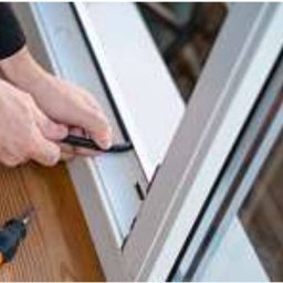 Serwis Domowy Valdi , Waldemar Michalski - Montaż uszczelki w oknie PCV. Dłonie aplikują czarną gumową uszczelkę w ramę okienną. Obok leży śrubokręt z pomarańczową rączką. Widok z bliska na drewnianą podłogę.