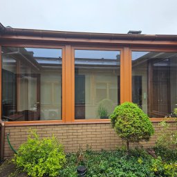 Serwis Domowy Valdi , Waldemar Michalski - Zewnętrzny widok przeszklonej werandy z brązowymi ramami okiennymi i ceglaną podstawą, otoczonej zielenią. Widoczne odbicia w szybach.