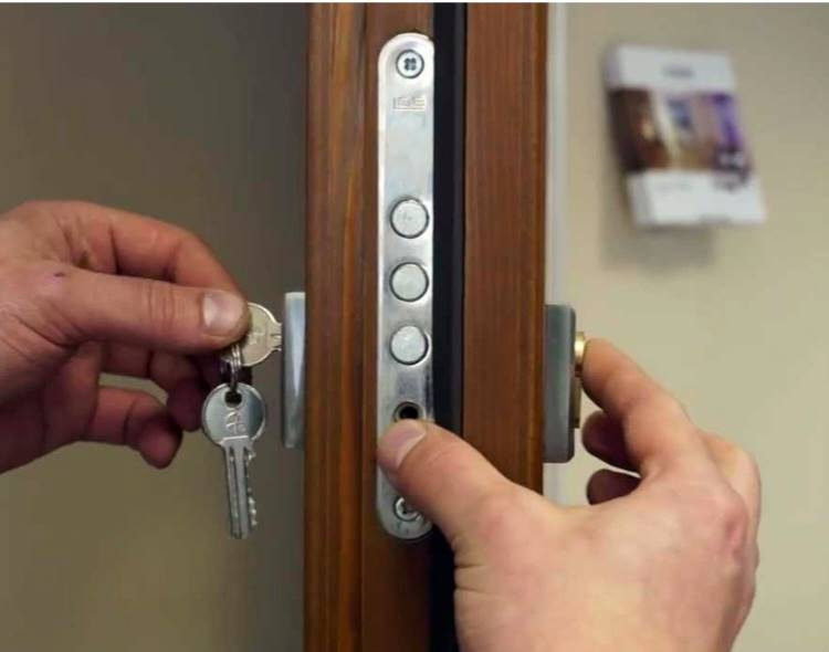 Otwieranie brązowych drzwi wejściowych z zamkiem wielopunktowym za pomocą pęku kluczy, z widocznym plakatem na ścianie w tle.