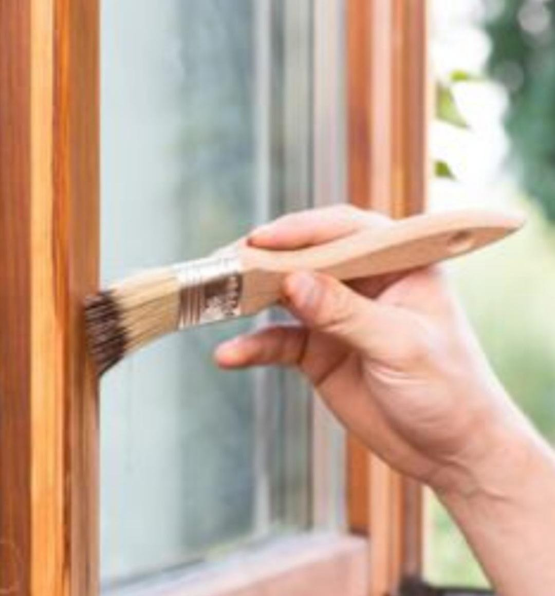 Malowanie drewnianej ramy okiennej pędzlem z brązową farbą, widoczny fragment szyby i tło z zielenią.