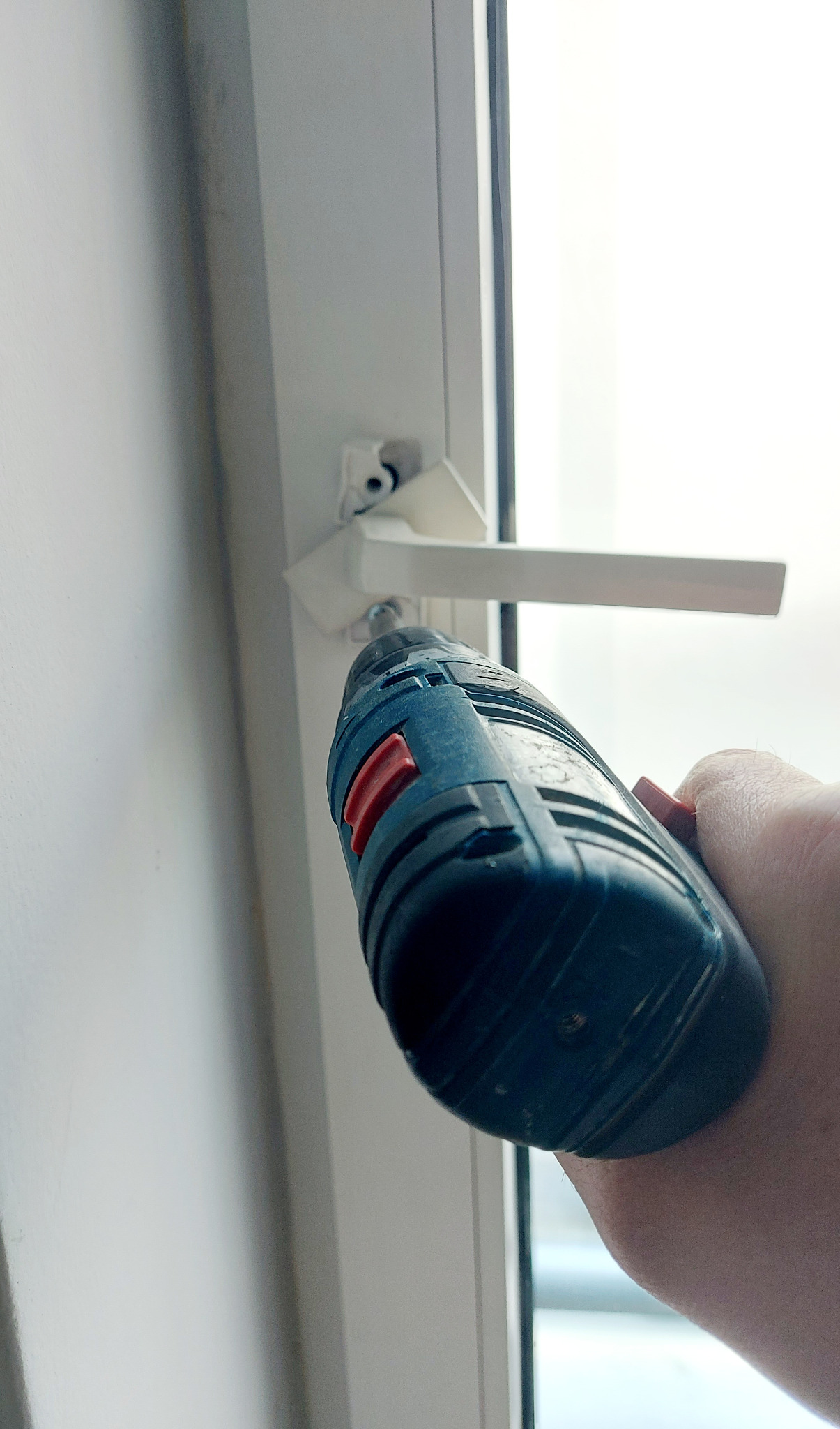 Przykręcanie białej klamki okiennej do ramy okna PCV za pomocą wkrętarki akumulatorowej.