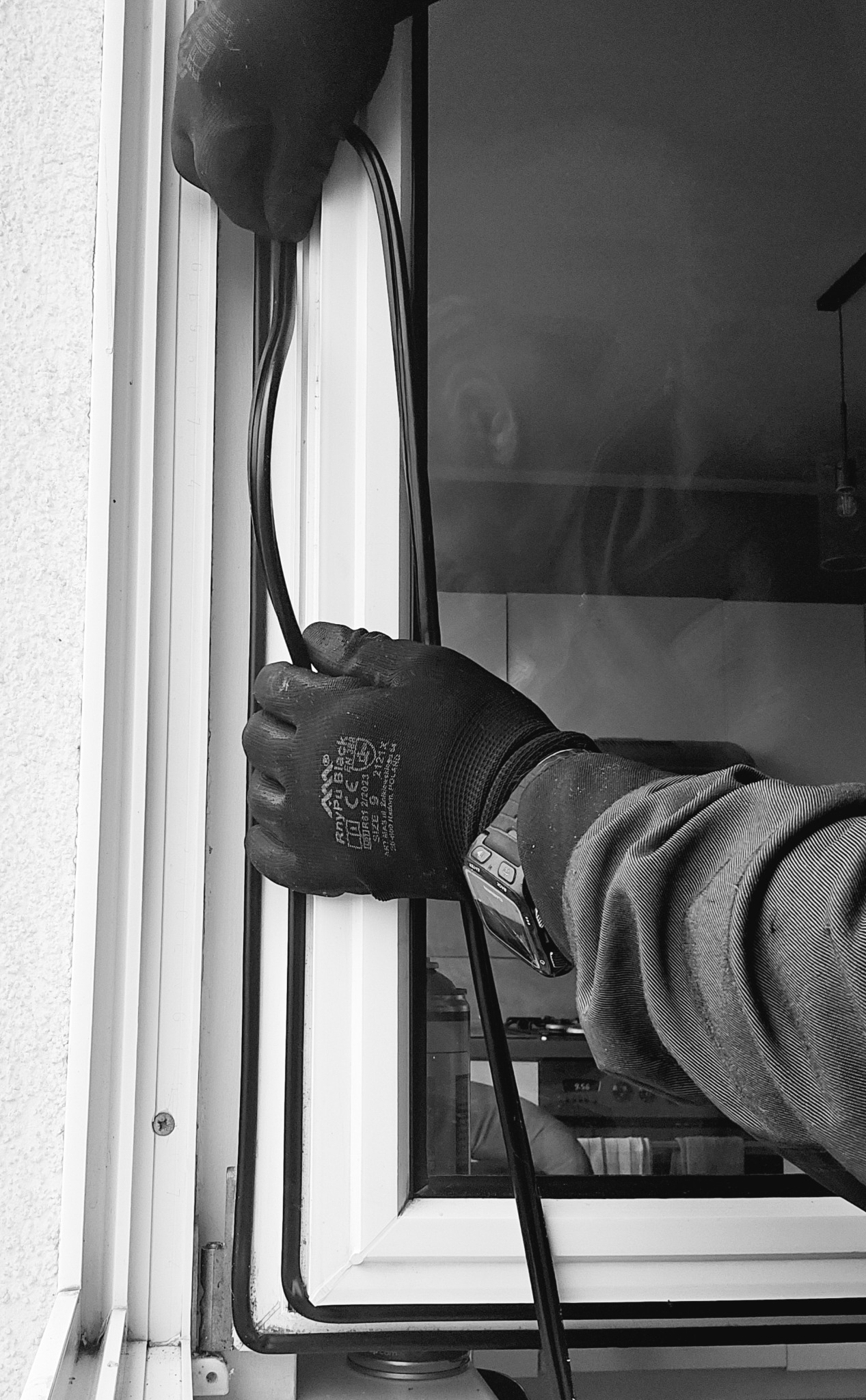 Czarno-białe zdjęcie dłoni w czarnej rękawicy roboczej, instalującej czarną uszczelkę w białej ramie okiennej; widoczny fragment ściany zewnętrznej i odbicie wnętrza pomieszczenia w szybie.