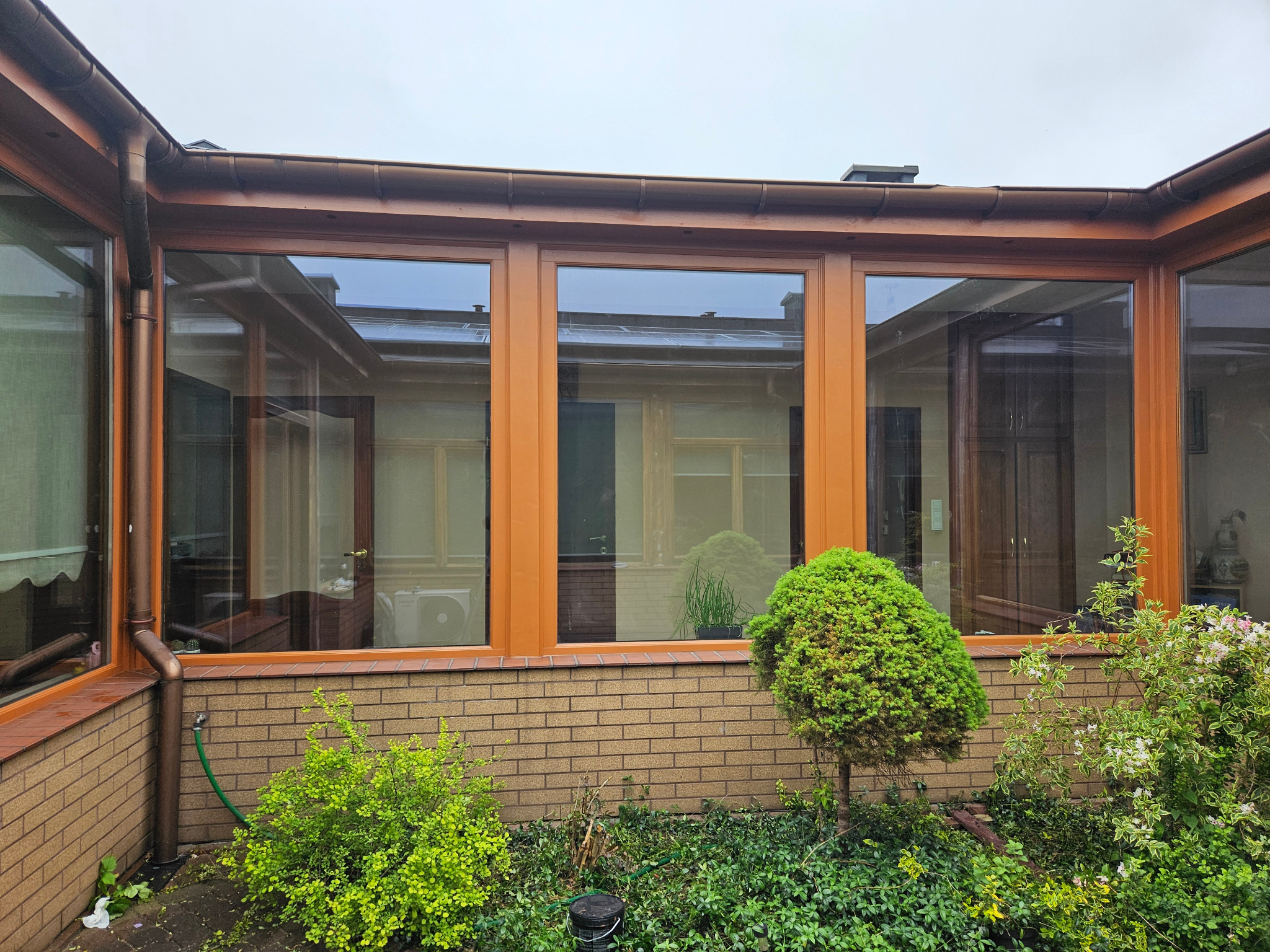 Zewnętrzny widok przeszklonej werandy z brązowymi ramami okiennymi i ceglaną podstawą, otoczonej zielenią. Widoczne odbicia w szybach.