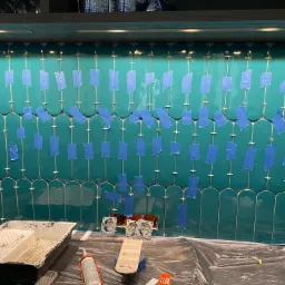 Montaż zielonych, podłużnych płytek na ścianie, zabezpieczonych niebieską taśmą malarską i krzyżykami dystansowymi; widoczne narzędzia glazurnicze i puszki elektryczne.