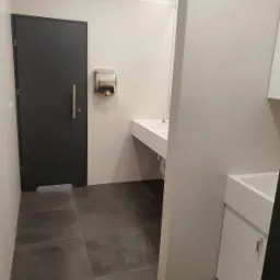 Nowoczesna łazienka publiczna z ciemną podłogą, białą umywalką, stalowym suszarką do rąk i ciemnymi drzwiami.