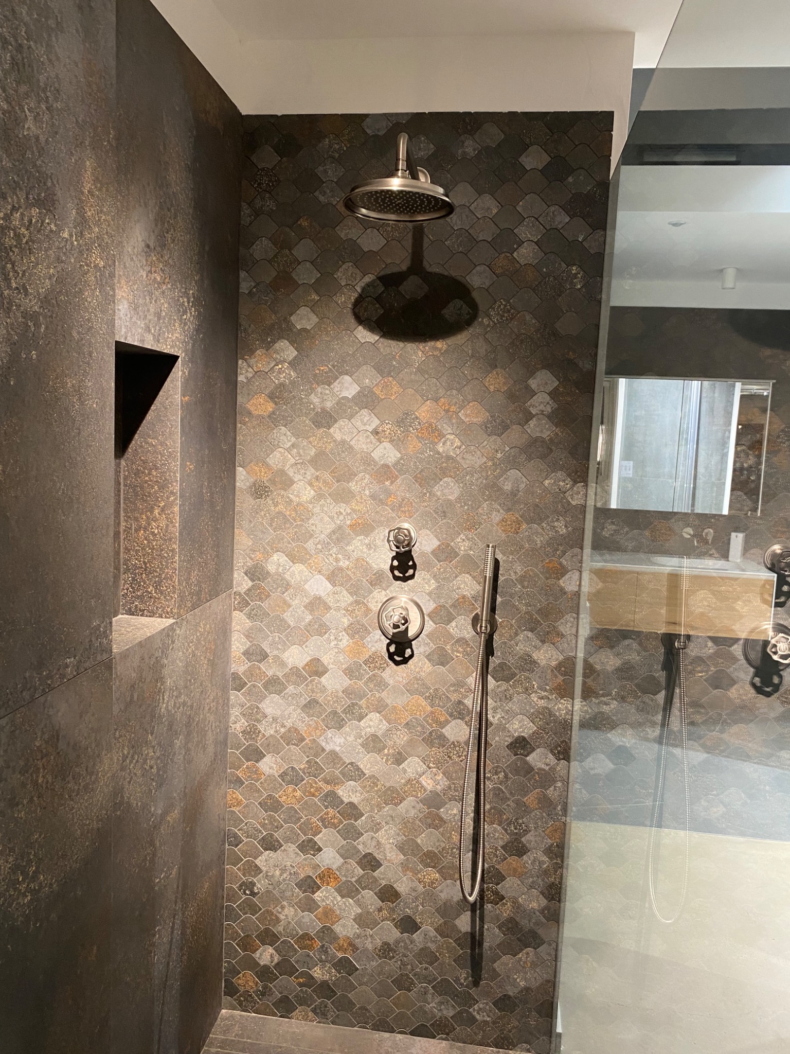 Kabina prysznicowa z ciemnymi, mozaikowymi płytkami w kształcie rybiej łuski, deszczownicą i wnęką w ścianie wykończoną ciemnym gresem.