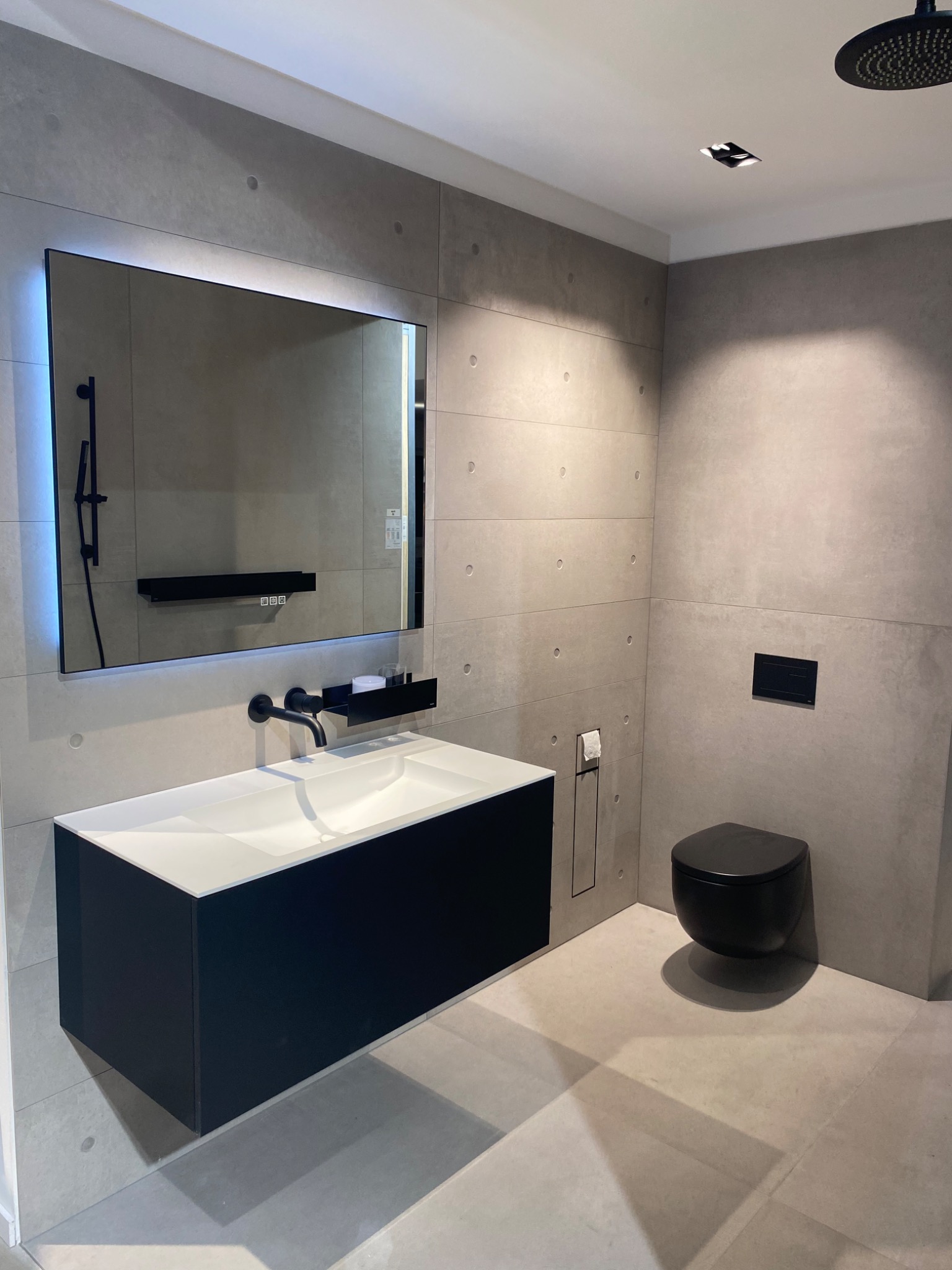 Nowoczesna łazienka z szarymi płytkami imitującymi beton, czarną armaturą i podświetlanym lustrem, wisząca toaleta w kolorze czarnym.