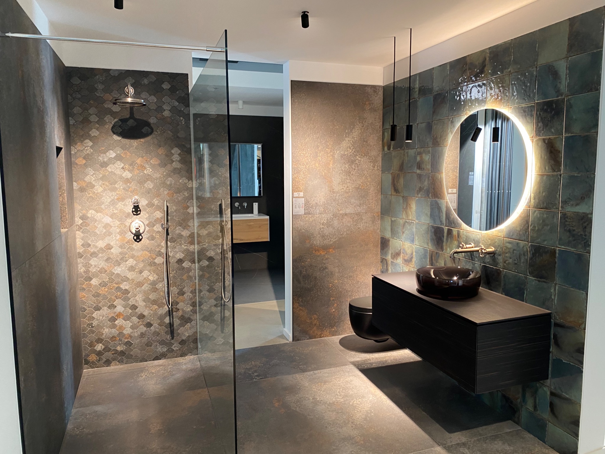 Nowoczesna łazienka z kabiną prysznicową typu walk-in, umywalką nablatową na wiszącej szafce z ciemnego drewna i okrągłym lustrem z podświetleniem LED, wykończona płytkami w odcieniach szarości...