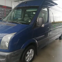 Niebieski bus myty w samoobsługowej myjni, widoczne krople wody na karoserii i mokra posadzka, w tle tablica z programami mycia.