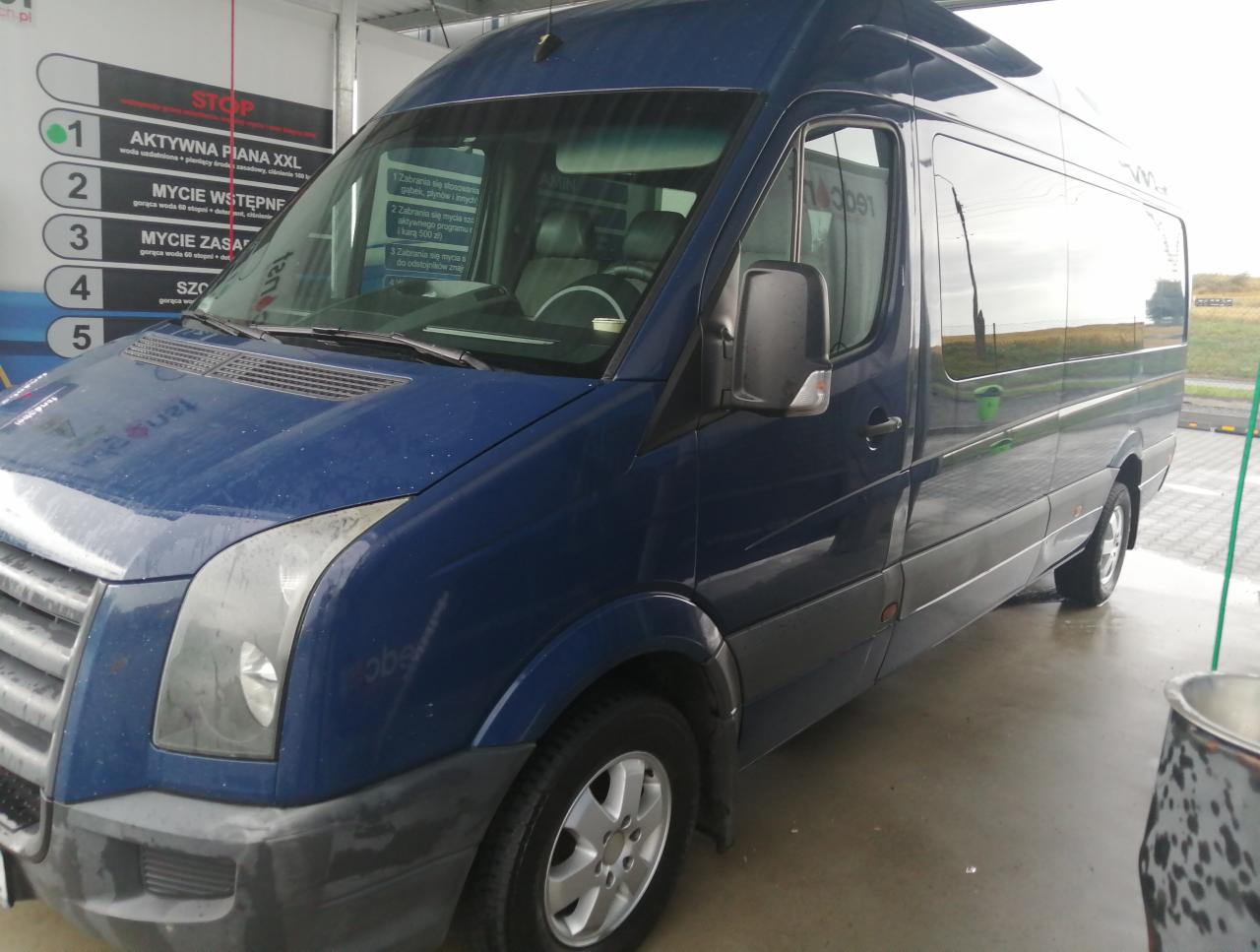 Niebieski bus myty w samoobsługowej myjni, widoczne krople wody na karoserii i mokra posadzka, w tle tablica z programami mycia.