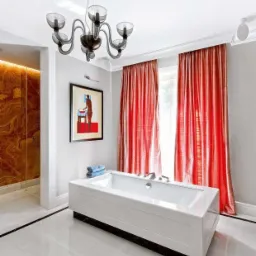 Nowoczesna łazienka z białą wanną obudowaną płytami, marmurową podłogą i ścianą prysznica z onyksu, oświetlona designerskim żyrandolem.