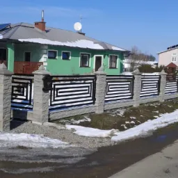 Nowoczesne ogrodzenie z elementami betonowymi i metalowymi, z geometrycznym wzorem, oddzielające posesję od ulicy, w tle zielony dom z panelami słonecznymi na dachu, resztki śniegu na trawniku.