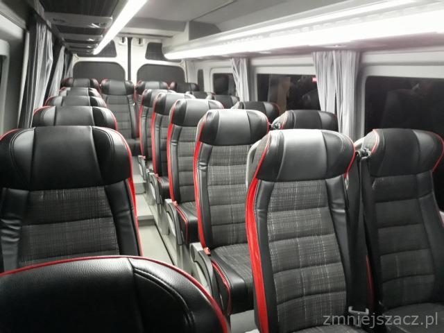 Wnętrze autobusu z czarnymi fotelami z czerwonymi akcentami i szarą tapicerką w kratkę, oświetlenie sufitowe.