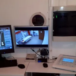 Stanowisko pracy z dwoma monitorami: na jednym wyświetlony obraz RTG dłoni, na drugim podgląd z kamery w pomieszczeniu z aparaturą medyczną i stołem do badań.