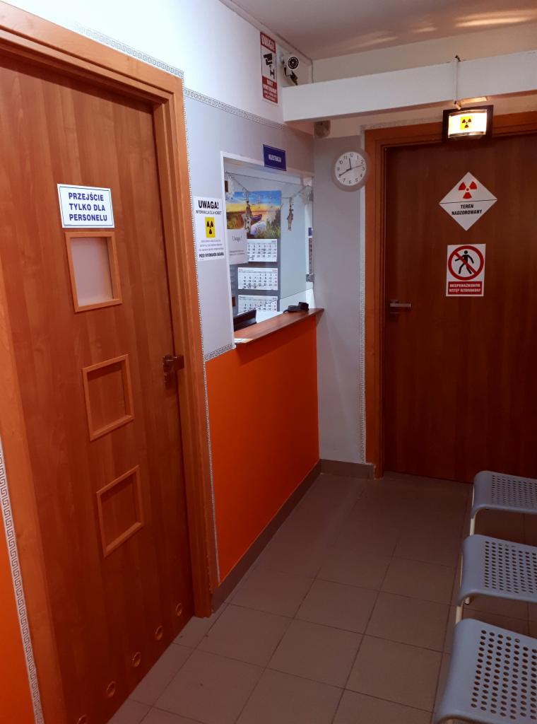 Poczekalnia w placówce medycznej: pomarańczowa lada recepcyjna, drzwi z napisem 'Przejście tylko dla personelu', drzwi z symbolem ostrzegawczym przed promieniowaniem, zegar na ścianie.