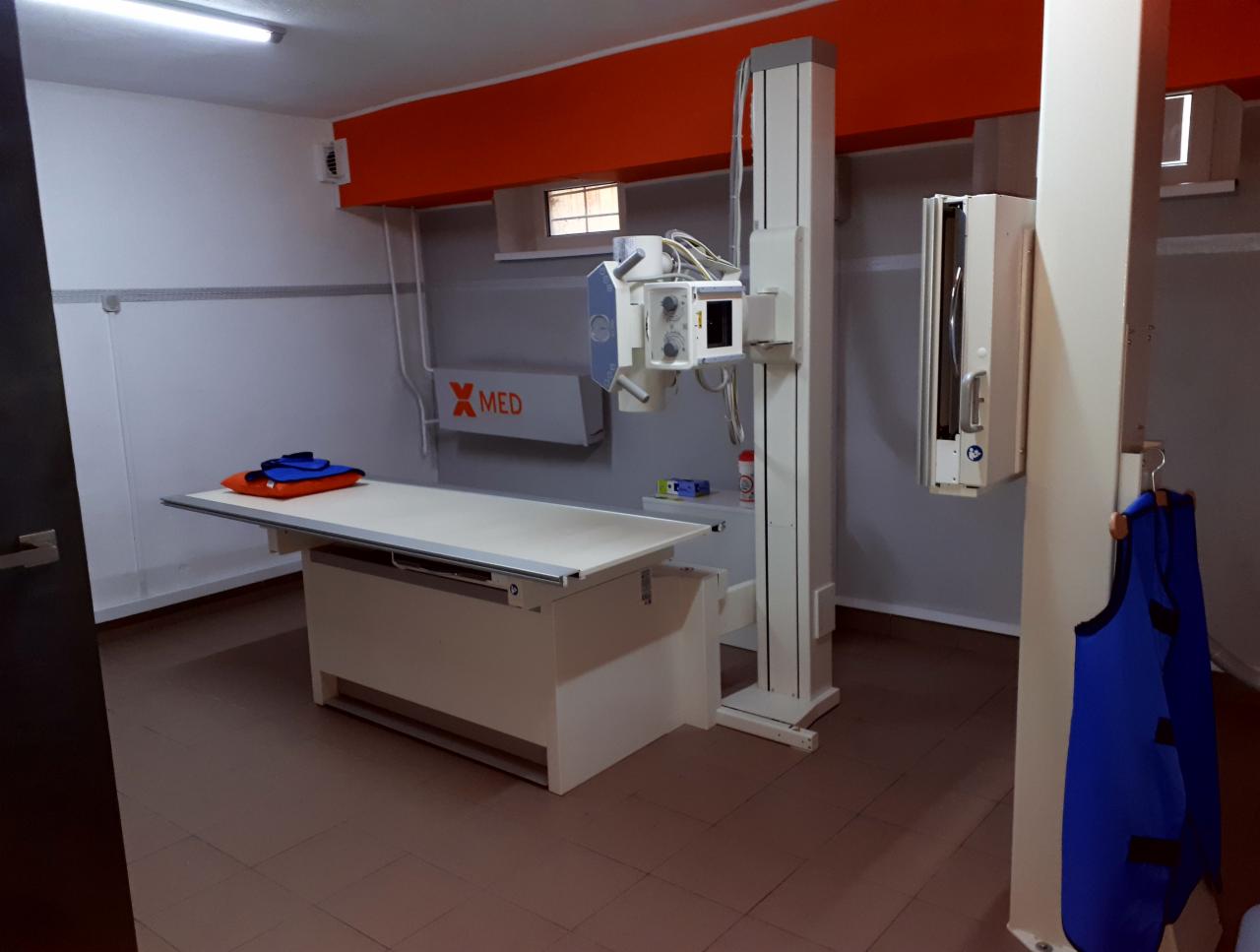 Pokój z aparaturą rentgenowską, na stole leżą ochronne fartuchy, ściany w kolorze szarym i pomarańczowym.