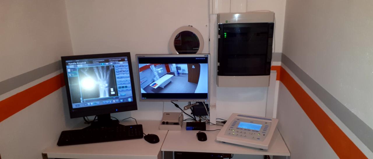 Stanowisko pracy z dwoma monitorami: na jednym wyświetlony obraz RTG dłoni, na drugim podgląd z kamery w pomieszczeniu z aparaturą medyczną i stołem do badań.