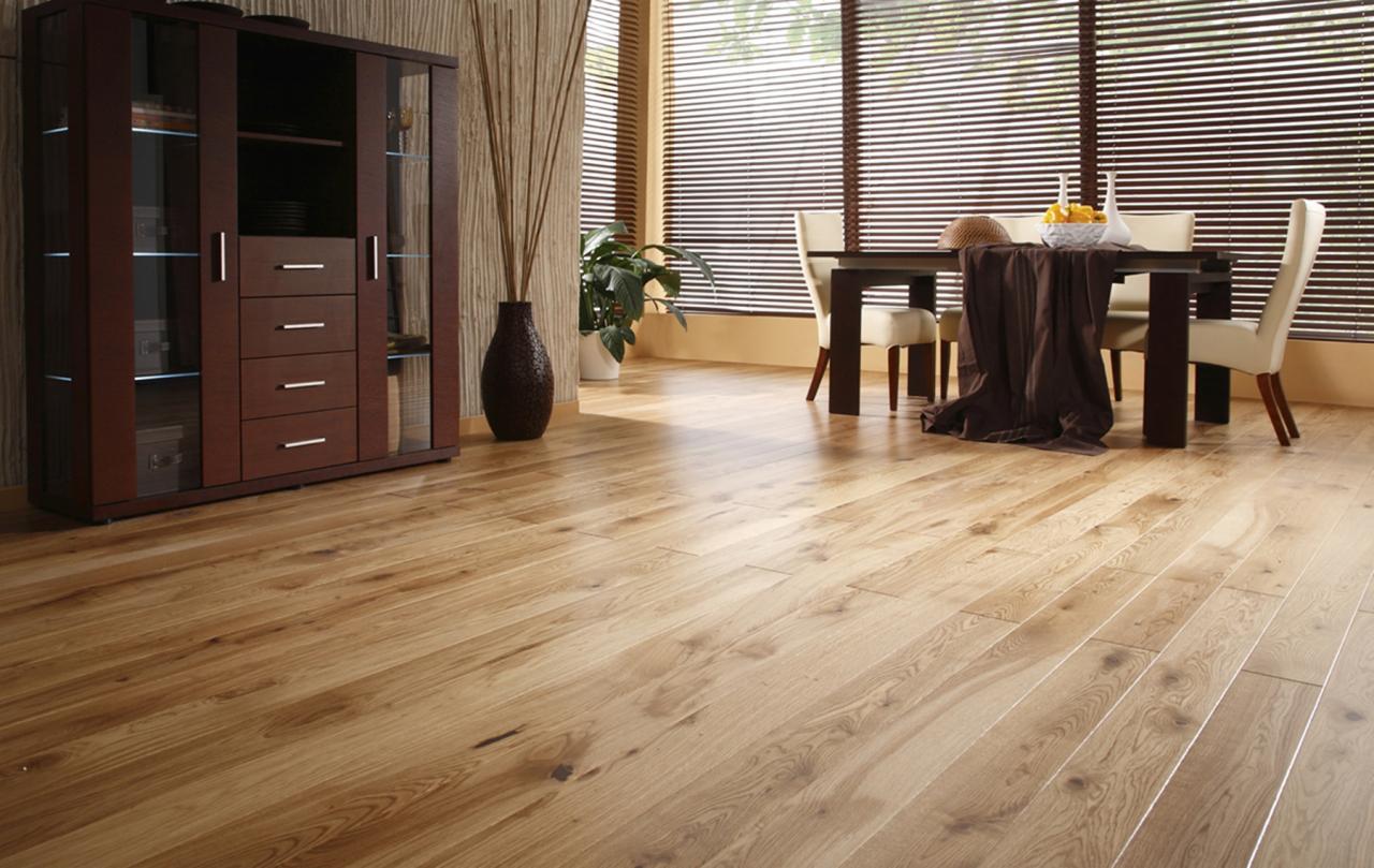 Jasna, drewniana podłoga z widocznym usłojeniem, w tle stół z krzesłami, zasłonięte żaluzjami okno, brązowa komoda z przeszkleniami oraz wazon z suchymi gałązkami.