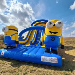 Klaudiusz Góra - Nadmuchiwania zjeżdżalnia w stylu Minionków na trawie, błękitne niebo w tle. Żółto-niebieska konstrukcja z dwoma postaciami Minionków po bokach.
