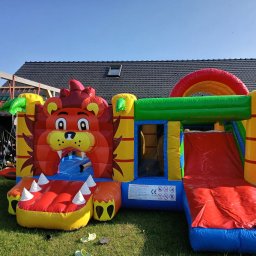 Klaudiusz Góra - Kolorowy dmuchany zamek w kształcie lwa z tunelem i zjeżdżalnią, ustawiony na trawie przed domem z ciemnym dachem. Widoczne zasady bezpieczeństwa.