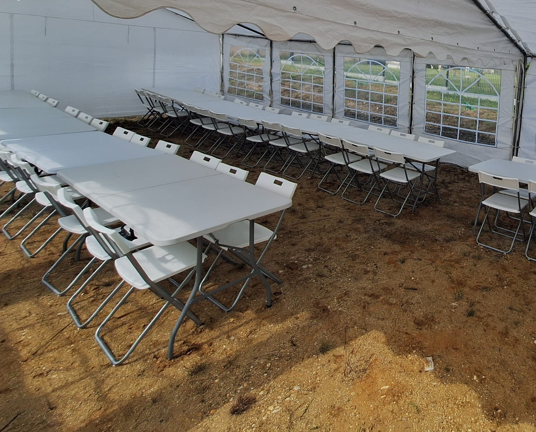 Biały namiot imprezowy z rzędami stołów i krzeseł. Widok na zewnątrz przez okna z kratownicą. Podłoże ziemne. Przygotowanie do wydarzenia.