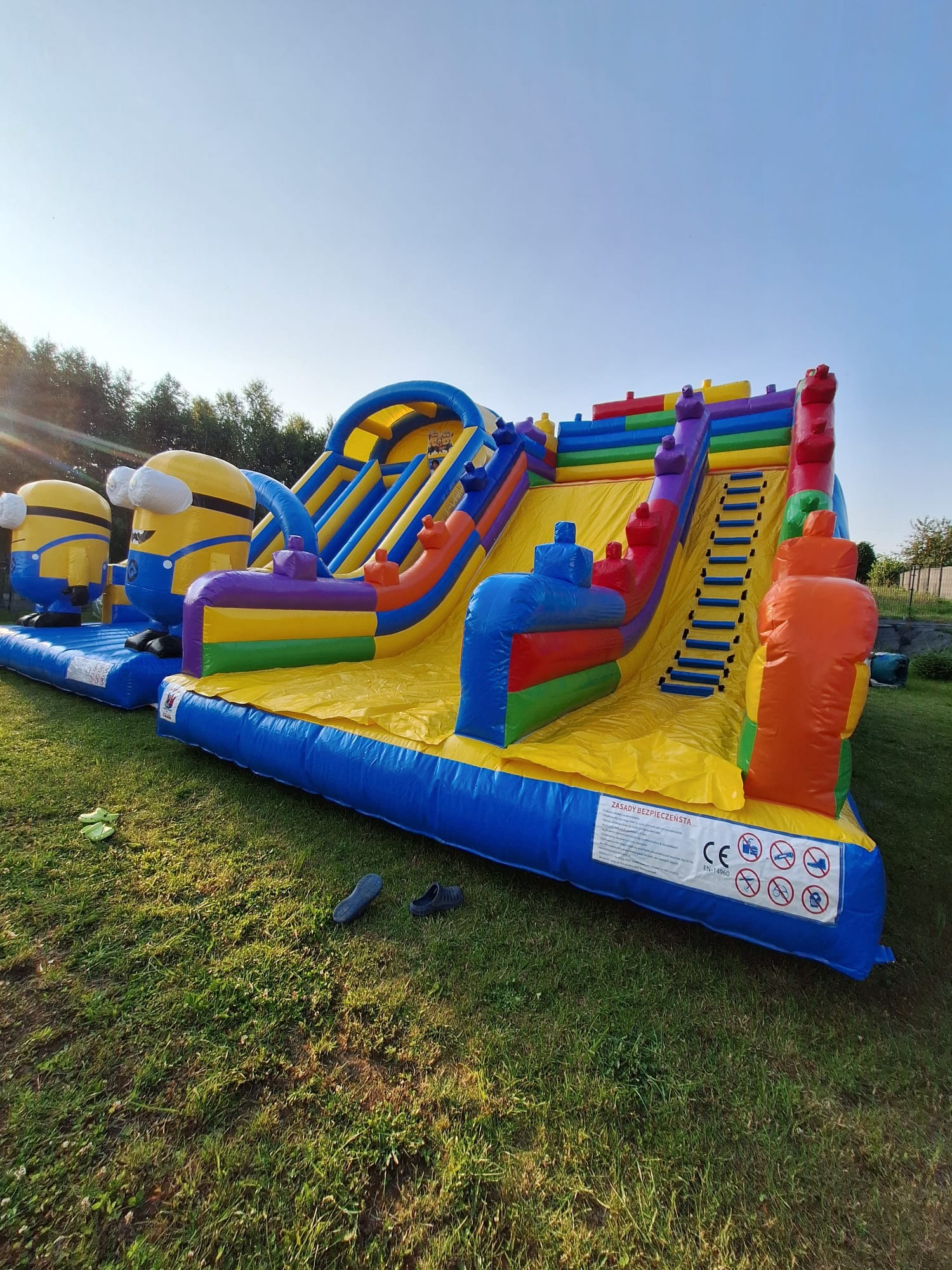Kolorowy dmuchany zamek ze zjeżdżalnią i motywem Minionków na trawie, w tle drzewa i błękitne niebo. Widoczne zasady bezpieczeństwa na zamku.