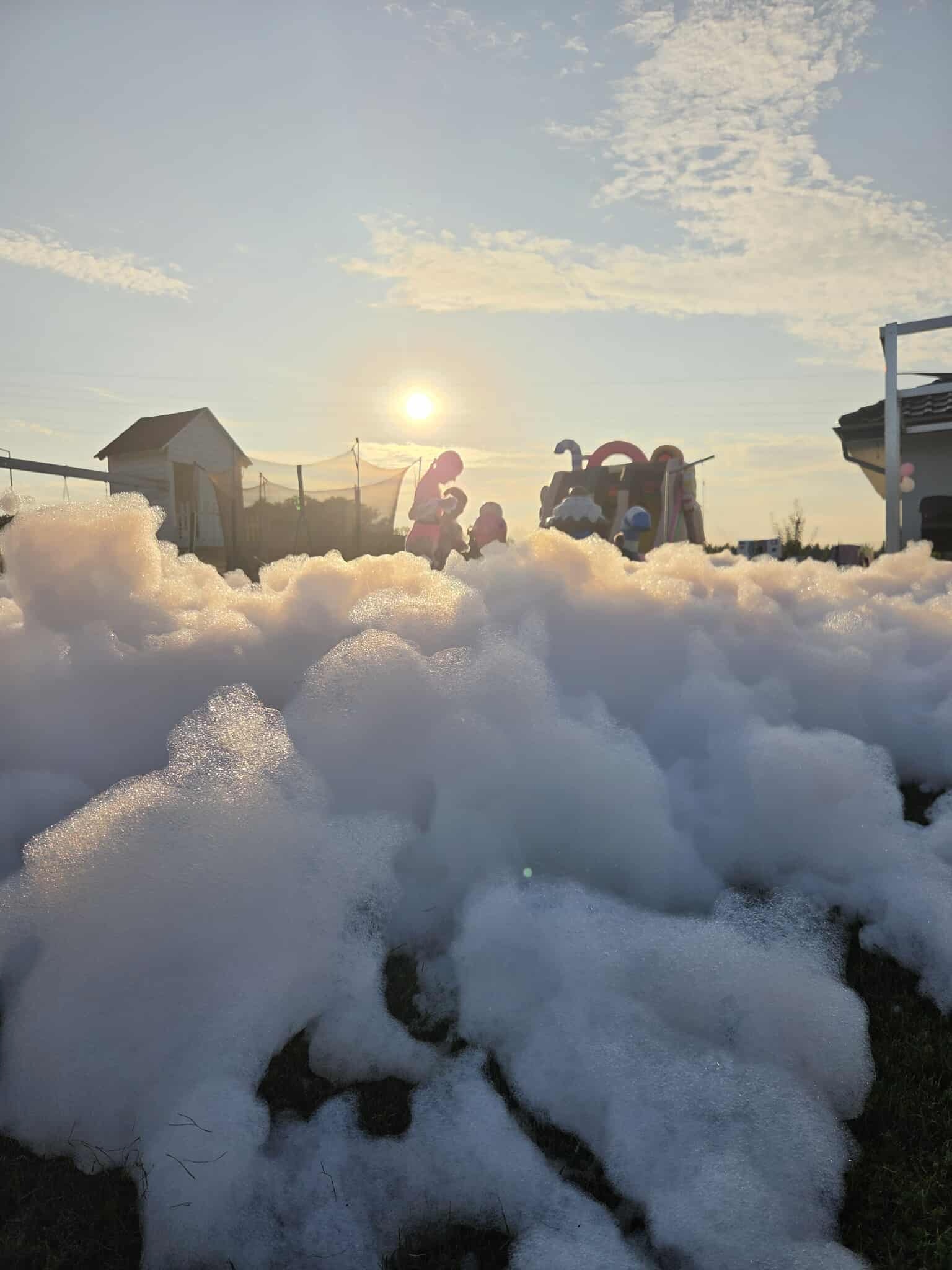 Piana party na trawie w promieniach zachodzącego słońca. W tle dmuchany zamek i bawiące się dzieci. Unikalne ujęcie z poziomu gruntu.