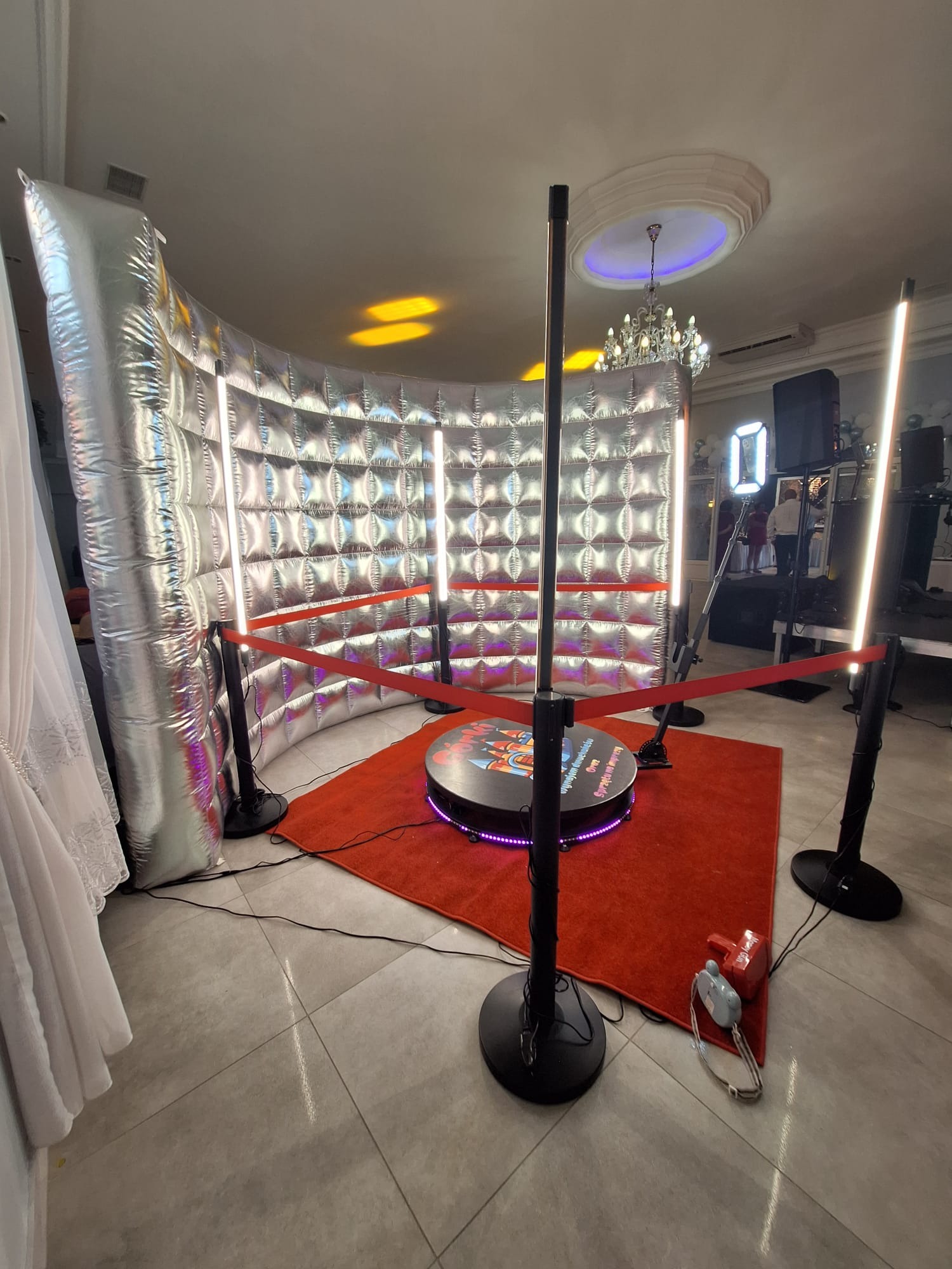 Kompletna, srebrna fotobudka 360 z oświetleniem LED i czerwonym dywanem, gotowa do użycia na imprezie. Widoczny dmuchany, srebrny łuk i dekoracyjne światła.