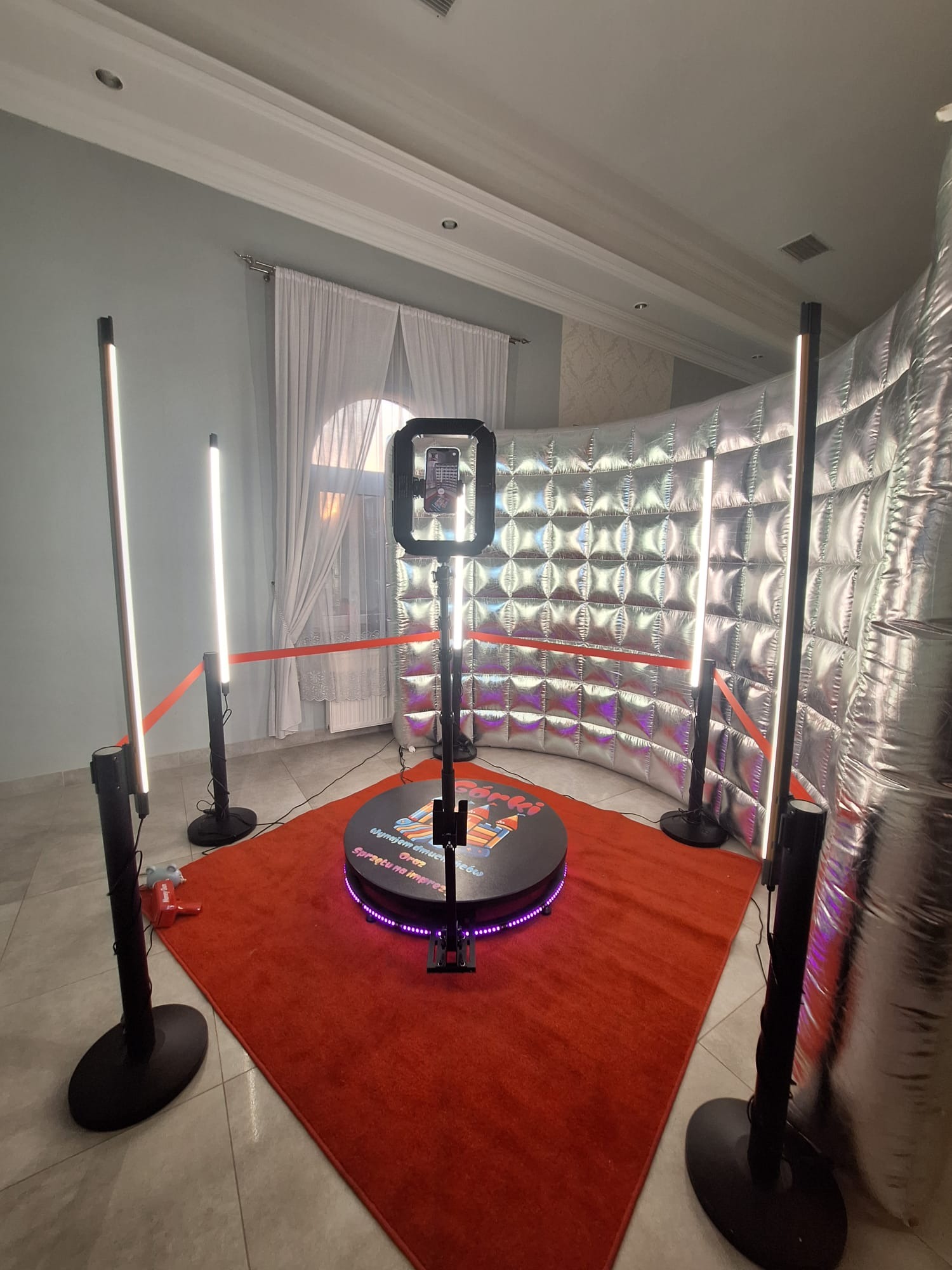 Kompletna, gotowa do użycia fotobudka 360 z oświetleniem LED i srebrnym tłem, ustawiona na czerwonym dywanie w jasnym pomieszczeniu. Widoczne elementy dekoracyjne.