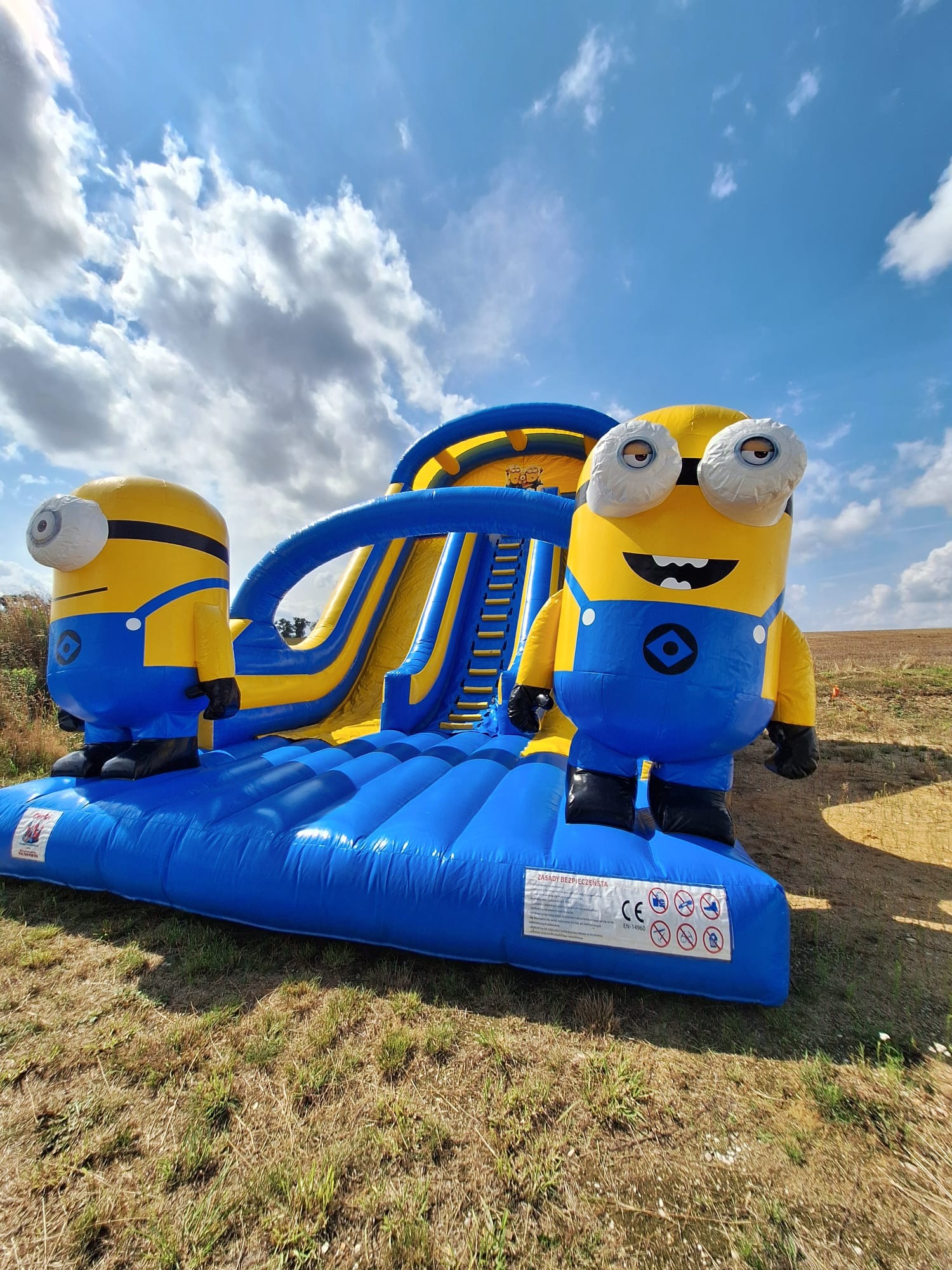 Nadmuchiwania zjeżdżalnia w stylu Minionków na trawie, błękitne niebo w tle. Żółto-niebieska konstrukcja z dwoma postaciami Minionków po bokach.