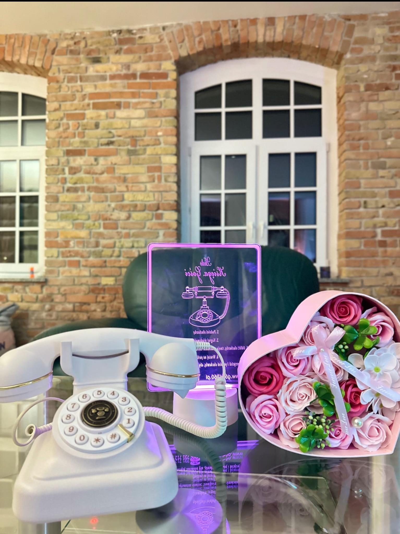Dekoracyjny, biały telefon retro obok pudełka w kształcie serca z różami i podświetlanej tabliczki z rysunkiem telefonu na tle ceglanej ściany z oknami.