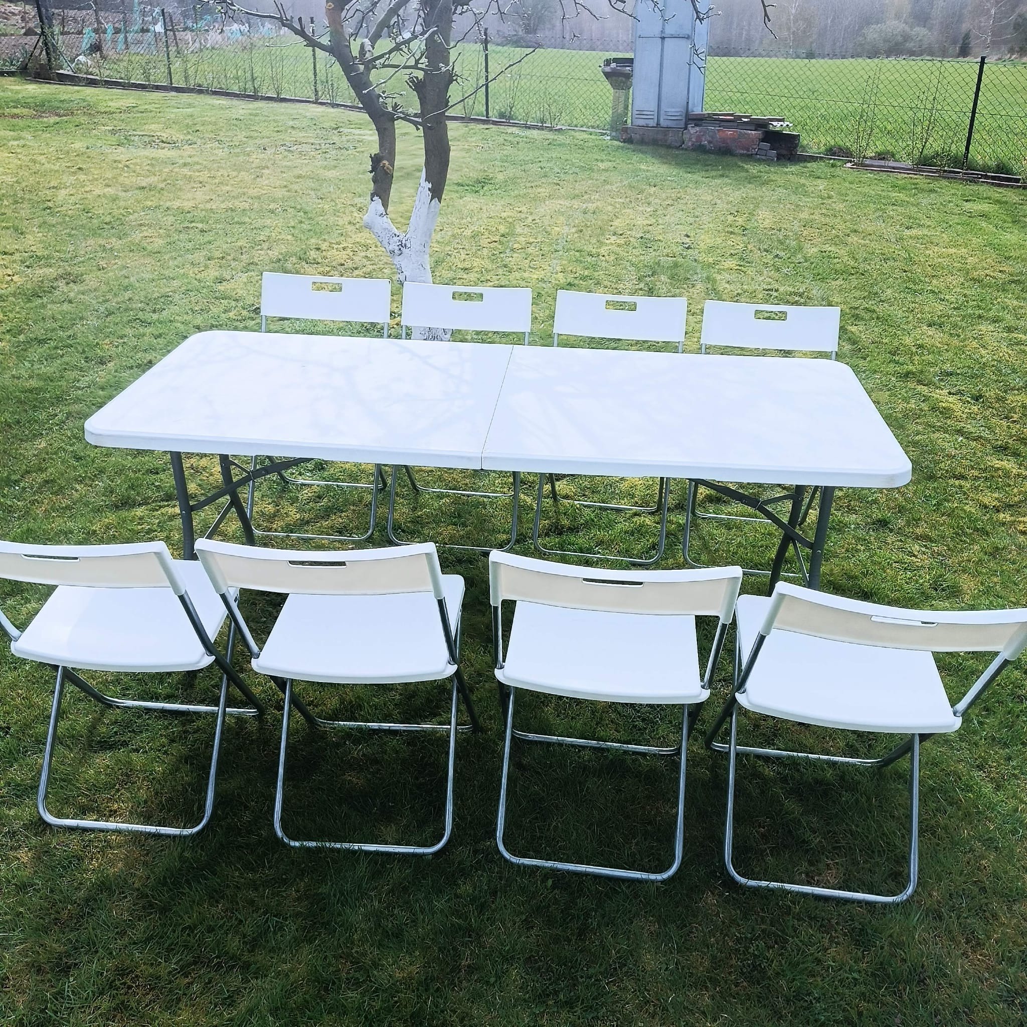 Biały stół cateringowy z krzesłami na trawie, przygotowany do imprezy plenerowej. W tle drzewo i ogrodzenie.