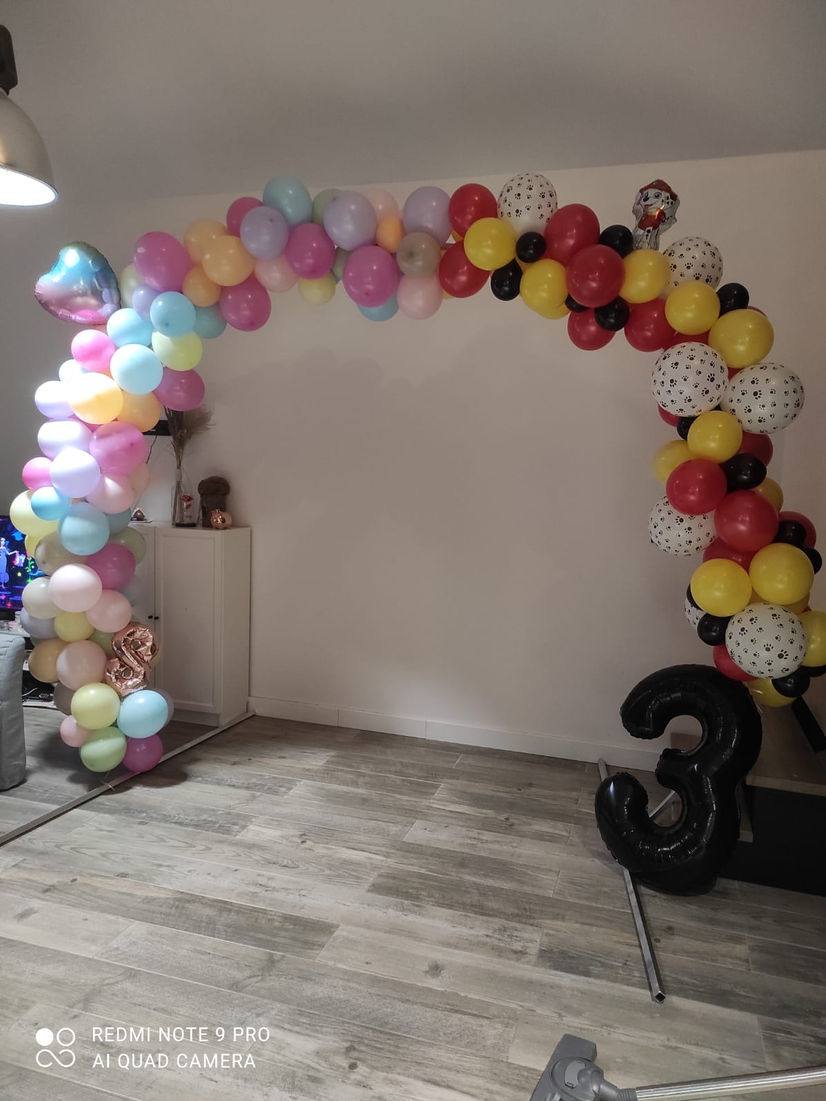 Aranżacja balonowa w kształcie łuku, z balonami w pastelowych kolorach i balonem w kształcie cyfry 3, ustawiona w pomieszczeniu na drewnianej podłodze.