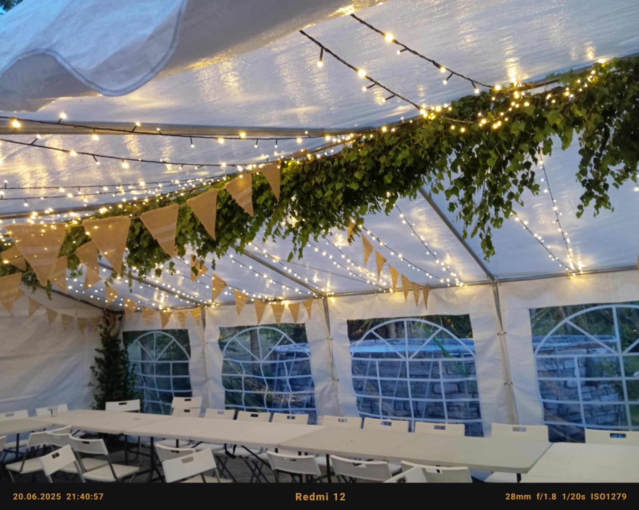 Udekorowany namiot imprezowy z girlandami, liśćmi i trójkątnymi proporczykami. Wnętrze z białymi stołami i krzesłami, widok na zewnątrz przez okna.