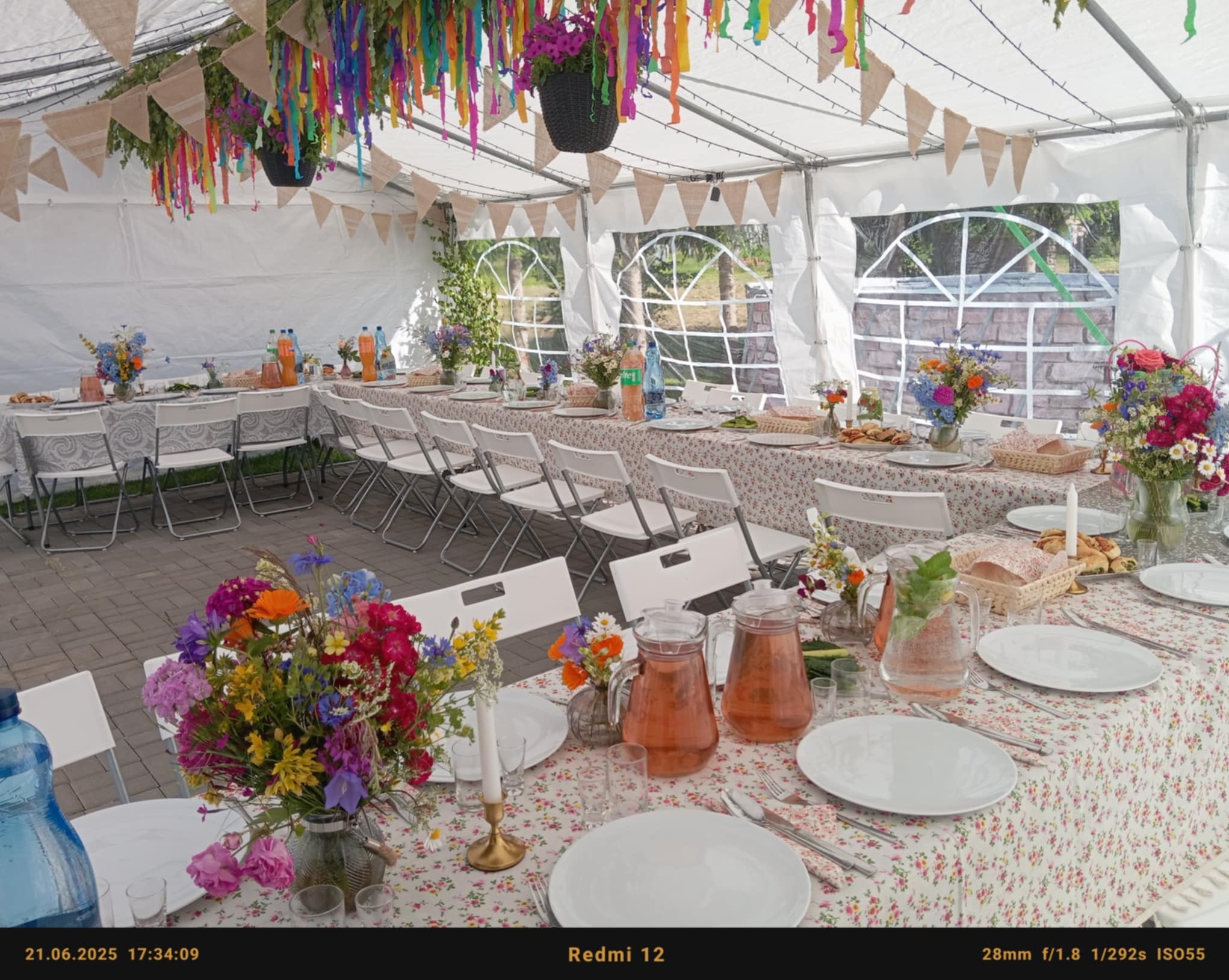 Udekorowany stół pod namiotem z kwiatami, zastawą i napojami. Dekoracje z kolorowych wstążek i proporczyków. Przygotowania do uroczystości.