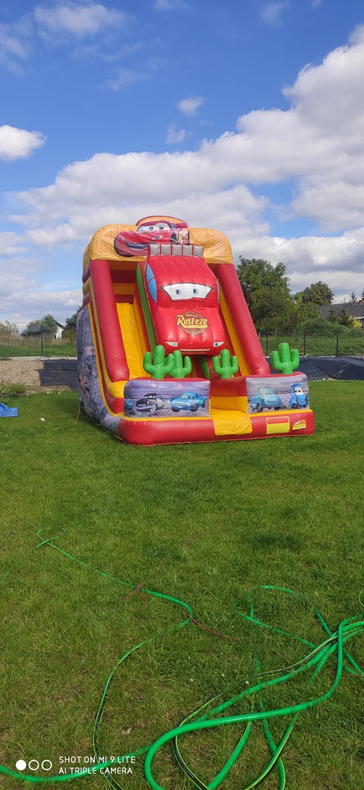 Kolorowa dmuchana zjeżdżalnia z motywem 'Auta' na trawie, w tle drzewa i błękitne niebo z chmurami. Widoczne detale: kaktusy i postacie z bajki.