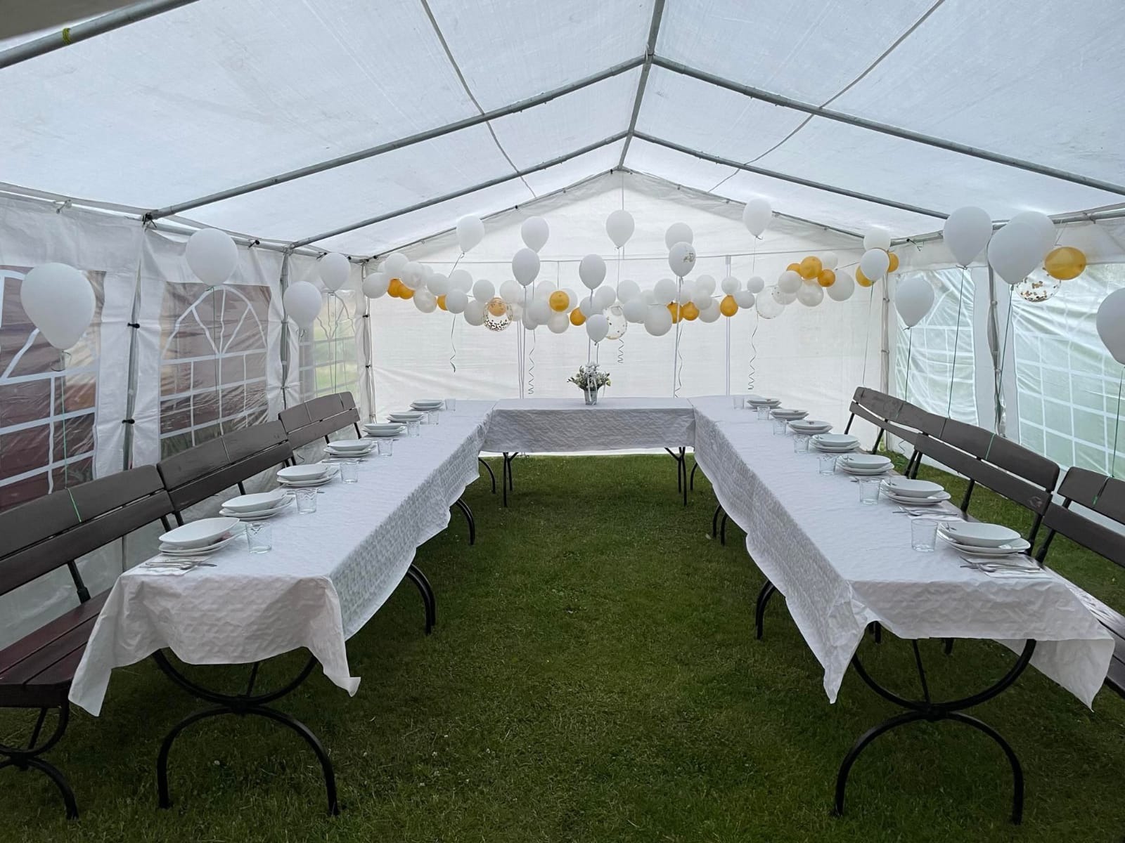 Udekorowany namiot na imprezę: białe i złote balony, stoły w kształcie litery U z białymi obrusami i nakryciami, ławki dla gości. Przygotowania do przyjęcia.