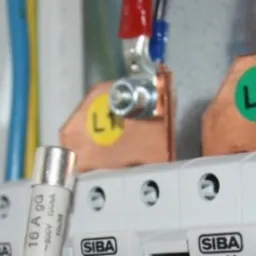 Szczegółowe ujęcie wnętrza skrzynki elektrycznej z widocznymi bezpiecznikami SIBA 10x38, przewodami oznaczonymi L1, L2 i L3 oraz elementami mocującymi.