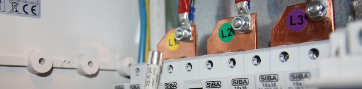 Szczegółowe ujęcie wnętrza skrzynki elektrycznej z widocznymi bezpiecznikami SIBA 10x38, przewodami oznaczonymi L1, L2 i L3 oraz elementami mocującymi.