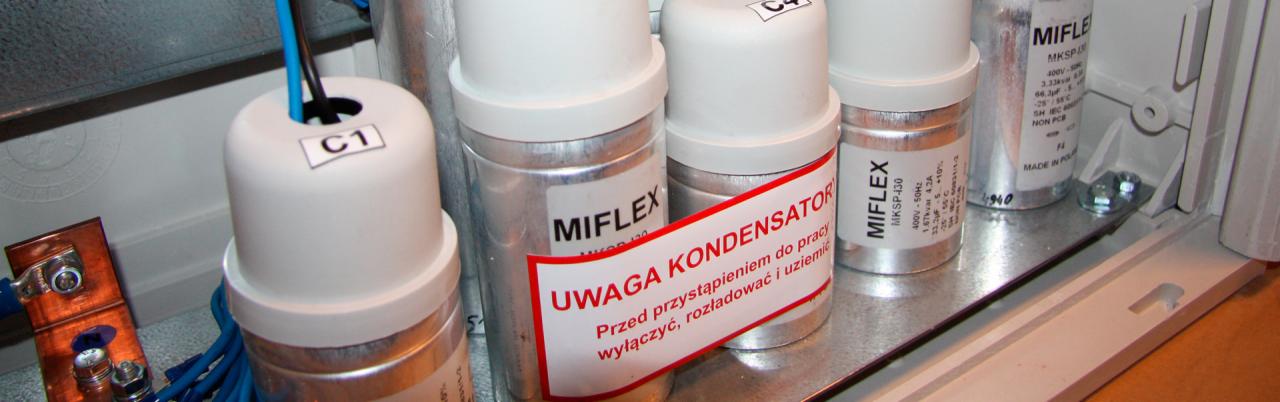 Szereg kondensatorów MIFLEX z ostrzeżeniem 'Uwaga Kondensator' wewnątrz białej skrzynki z widocznymi przewodami i elementami mocującymi.