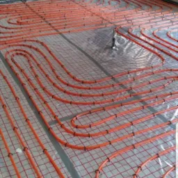 Pomarańczowe rury ogrzewania podłogowego ułożone na siatce w surowym wnętrzu, widok z góry, etap prac przed wylaniem posadzki. Fragment okna w tle.
