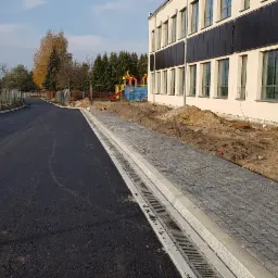 Nowa nawierzchnia asfaltowa i chodnik z kostki brukowej przed budynkiem, widoczne studzienki kanalizacyjne i przygotowany teren pod trawnik