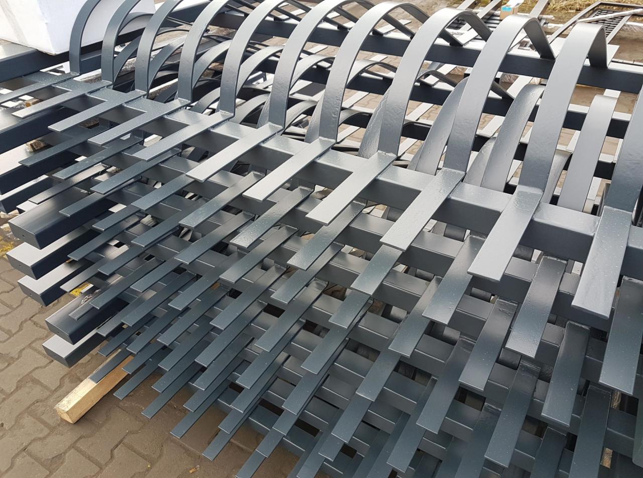 Stos pomalowanych na szaro elementów metalowych, prawdopodobnie ogrodzenia, z charakterystycznymi łukowymi zakończeniami, ułożonych warstwami na palecie.