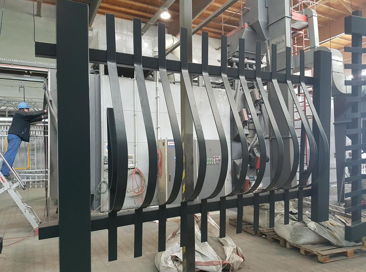 Metalowa barierka po malowaniu proszkowym, w tle pracownik na drabinie i piec do malowania.