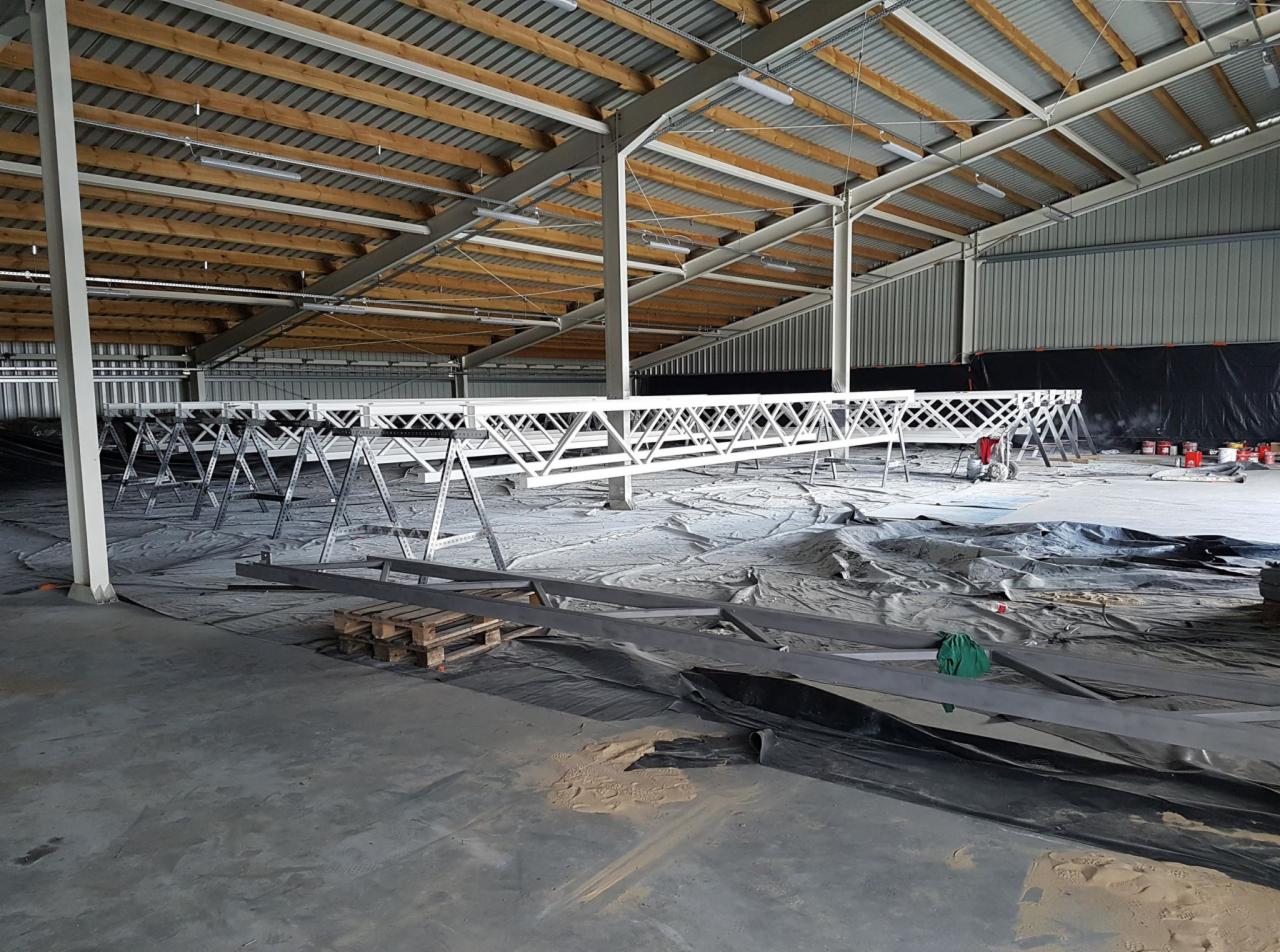 Biała, metalowa konstrukcja kratowa na podporach w hali, przykryta folią ochronną, widoczne puszki z farbą i paleta drewniana.