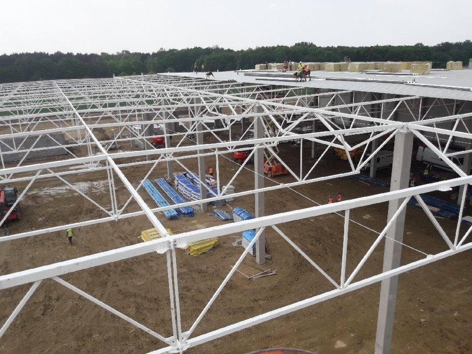 Konstrukcja stalowa hali przemysłowej z białymi elementami kratownicowymi, widok z góry na plac budowy z pracownikami na dachu i materiałami budowlanymi na ziemi.